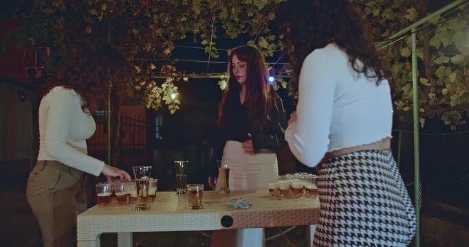 Three young women enjoy a beer pong game at a lively backyard party, illuminated by string lights, creating a fun and social atmosphere.