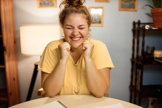 Indoor image of super excited female with hair bun doing winner gesture with clenched fists and closed eyes sitting at table at home, achieving long-term goal passing exam or getting new job