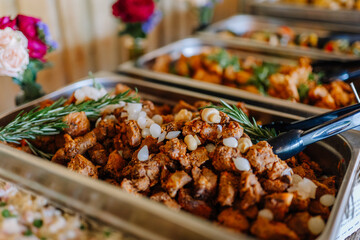 Close-up of a buffet tray filled with seasoned meat garnished with rosemary and pearl onions, served with tongs at a catered event.