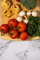 Fresh vegetables including tomatoes, mushrooms, broccoli, and kale, along with pasta, are arranged on a wooden cutting board.