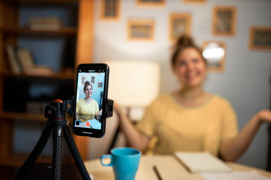 Selective focus of smartphone on tripod with image of Caucasian female English teacher recording video tutorial to get new followers and clients sitting in her cabinet at home gesticulating
