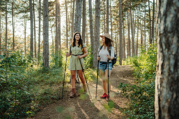 Two female friends hike or trek through scenic forest with hiking sticks and gear	
