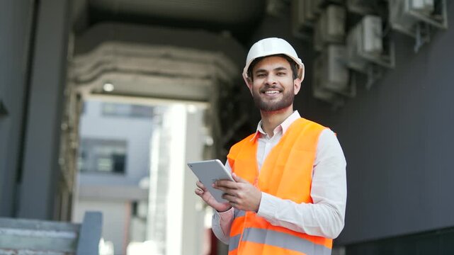 Portrait of smiling professional engineer wearing safety helme, vest standing at factory holding using digital tablet in hands. Headshot of confident handsome industry manager posing looking at camera - Powered by Adobe