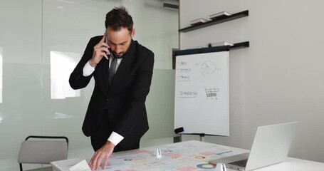 Serious Latin company executive man analyzing paper sales financial reports on workplace table, talking on mobile phone, discussing marketing research results with partner, coworker on cellphone - Powered by Adobe
