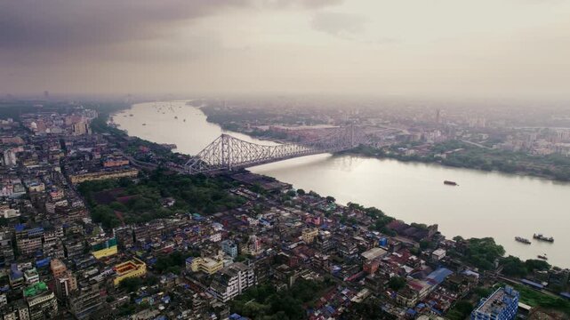 A cinematic daylight drone aerial shot of the historic heritage Howrah Bridge over holy Hooghly or Ganges river, surrounded by Kolkata or Calcutta city with a vibrant cloudy sky in the background.