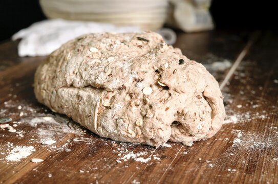 Close-up of raw artisan spelt bread dough with walnuts, carrots, oats, and seeds, resting on a floured wooden table before baking.