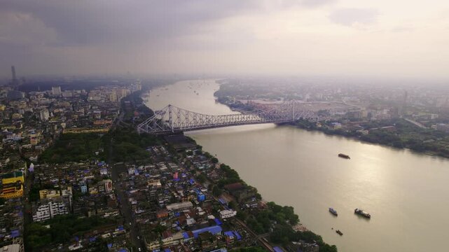 A cinematic daylight drone aerial shot of the historic or iconic Howrah Bridge over holy Hooghly or Ganges River, surrounded by Kolkata or Calcutta city with a vibrant cloudy sky in the background.