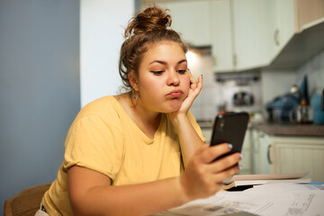 Annoyed female in home clothes pouting lips with frustration using smartphone to count tax index or using mobile application for online payment, paying for utility bills, having financial problems