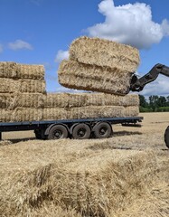 Special equipment loads large bales of straw onto a trailer. Work in the field after the harvest under the summer sky. The photo shows an important stage of agricultural work