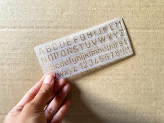 Hand holding a transparent alphabet plastic stencil against cardboard background.