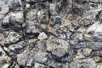 Rough Stone Wall or Rock Texture Background. Closeup, horizontal view of rugged, weathered rock face or stone wall with various shades of gray, white, and hint of green moss, showing natural cracks