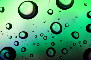 A macro shot of clear water droplets and bubbles on a colorful surface, reflecting light and creating a green and black abstract background.
