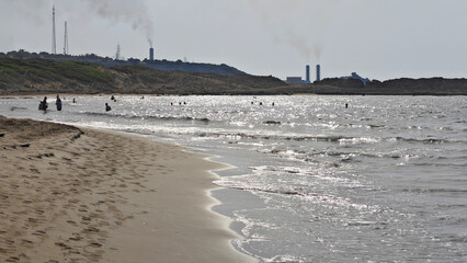 Sunlit Waves and Swimmers on a Cyprus Beach. Waves gently roll onto a sandy beach in Cyprus, with several people swimming in the sunlit water. Industrial chimneys are visible on the distant horizon.