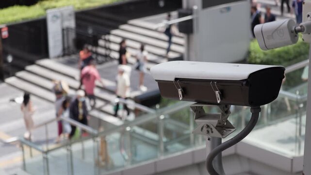 Urban security surveillance camera mounted in a bustling city area, capturing pedestrians navigating busy steps. Clear focus on camera with blurred background of lively street activity