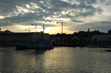 A boat on the water with cranes and buildings in the background at sunset.