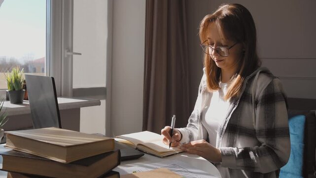 Young professional woman focused on working in home office taking notes in notebook while referring book, surrounded by laptop, study materials on organized desk with natural light penetrating inside