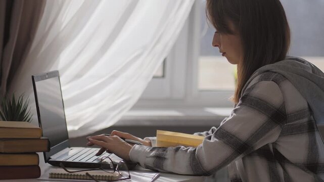 Natural sunlight filtering in through window, highlighting a focused student or woman freelancer working on a laptop taking handwritten notes surrounded by textbooks and glasses at her home workplace
