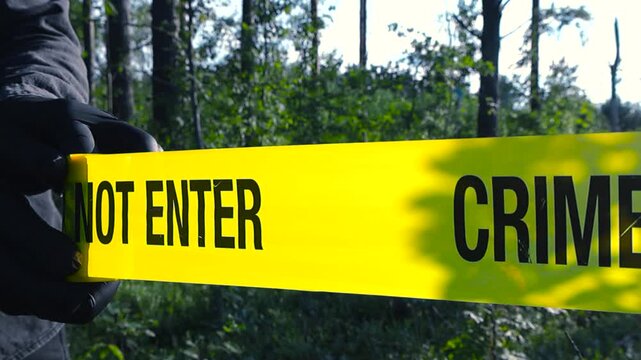 Yellow crime scene tape in close up being pulled in front of blurry wooded area with bushes. Forensic detective with black gloves, hands unrolling barricade tape, marking scene during sunny daylight