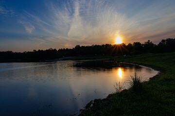 Golden sunset over a peaceful lake with shimmering reflections, grassy shoreline, and a dramatic sky filled with soft clouds, creating a serene and picturesque evening scene.