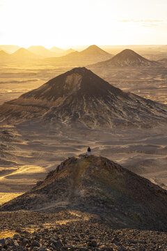 View of the sun-kissed desert landscape unfolds with layered mountains and a lone figure perched atop the foremost peak, Bawati, Giza Governorate, Egypt.