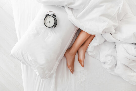 Womans legs peeking from under white bedding with alarm clock and pillow on bed - Powered by Adobe