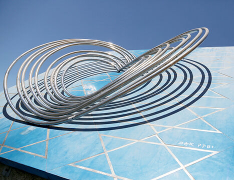 Contemporary metal sculpture on a vibrant blue wall in an urban outdoor setting under clear skies