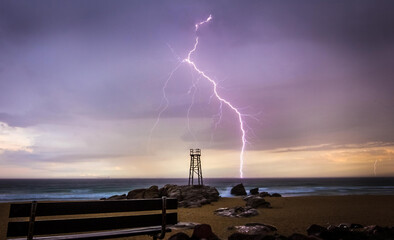 View of jagged lightning bolts dramatically illuminate the sky above a lifeguard tower on a rocky outcrop, contrasting with the calm beach, Redhead Beach, Newcastle, Australia.