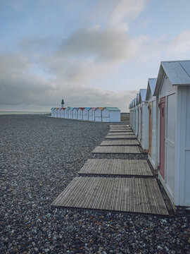 View of rows of quaint white beach huts with colorful accents line a pebble beach under a soft, diffused sky, casting long shadows, Le Tr&Atilde;&copy;port, Normandy, France.