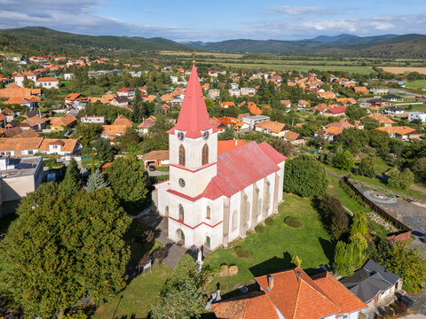 Aerial view of a church with a striking red roof stands proudly amidst verdant trees, overlooking the quaint village, Slovakia.