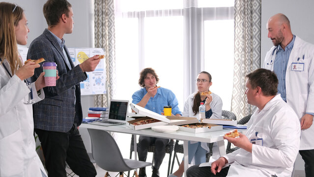 A group of successful doctors gathered around a table in a cozy clinic meeting room and are eating pizza. The hospital staff are dressed in white lab coats - Powered by Adobe