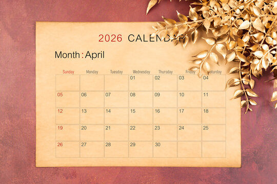 April 2026 calendar page with golden color leaves on red wooden background. - Powered by Adobe