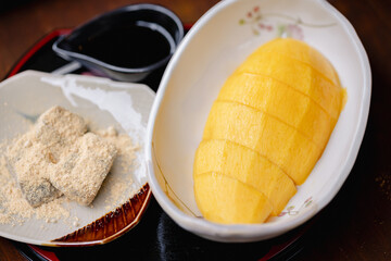 Sliced yellow fruit served on white plate with traditional Japanese style sweets coated in powder and small dish of dark sauce reflecting culture and culinary art