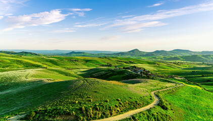 green flowering spring hills in countryside valley with rustic village road among blossoming grasslands leading far away to a sunset landscape in summer season