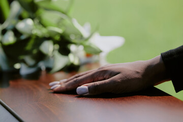 Black woman hand resting on wooden casket during funeral ceremony, blurred green background and floral arrangement visible, close up capturing gesture of farewell and mourning