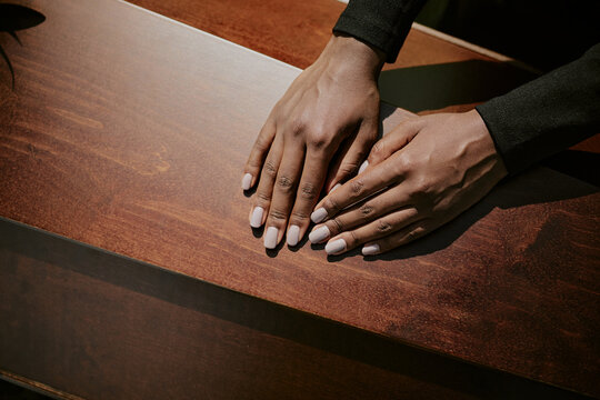 Black woman resting hands on wooden casket during funeral ceremony, fingers gently touching surface, showing grief and respect, close up of hands with natural light highlighting skin tone