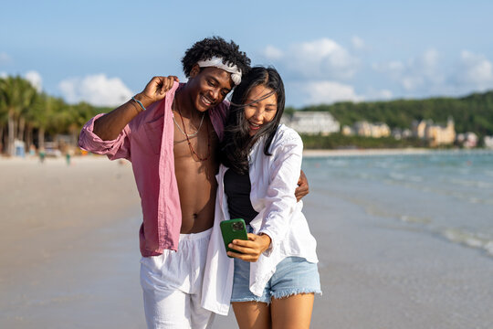 Diverse couple connected, embracing technology and social media while enjoying moments at the beach - Powered by Adobe