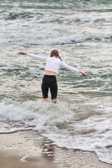 Fototapeta premium Young woman, brown hair, balances on baltic sea shoreline, embracing yoga posture. Overcast sky, gentle waves, evoke tranquility and focus. Natural, soothing setting for mindfulness