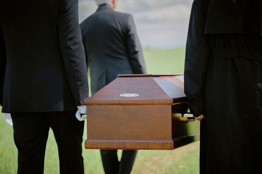 Caucasian middle aged men and woman carrying wooden coffin outdoors during funeral procession, walking together on grassy field with backs turned to camera, wearing formal attire