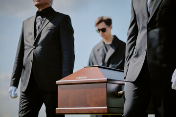 Caucasian middle aged men and young adult man wearing black suits carrying wooden coffin outdoors during funeral ceremony, young adult man wearing sunglasses walking behind casket