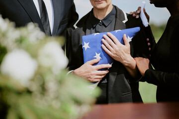 Middle aged Caucasian woman holding folded American flag while being comforted by Black woman and Caucasian man during funeral ceremony outdoors, hands visible, no faces shown