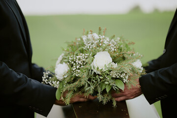 Two middle aged Caucasian men holding funeral casket adorned with floral arrangement outdoors, hands visible near bouquet, participating in burial ceremony