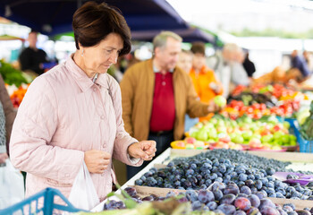 Positive mature woman in casual clothes picking fresh plums during shopping at grocery market