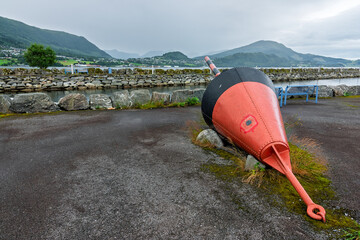 A vibrant orange and black buoy rests on a rocky surface near the coastline, framed by mountains and overcast skies.