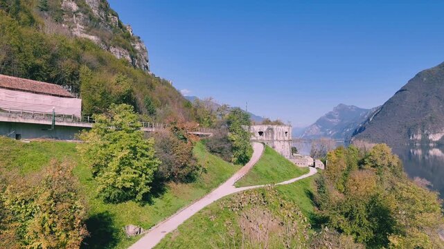 road next to lake Idro surrounded by mountains and old round stone building of Rocca d'Anfo in Italy