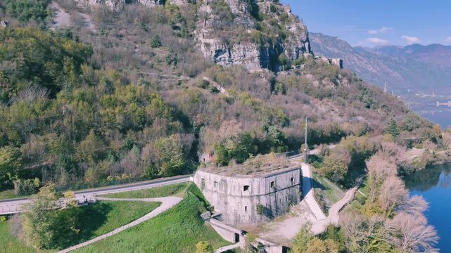 Rocca d'Anfo situated next to a road and lake Idro with a forested mountain under blue sky in Italy