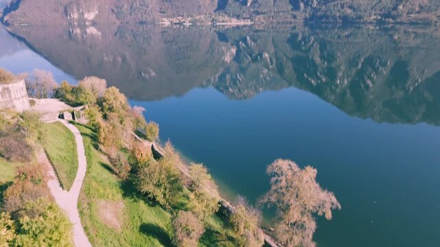 lake Idro reflecting green mountains, with Rocca d'Anfo on the left under a clear blue sky. Italy