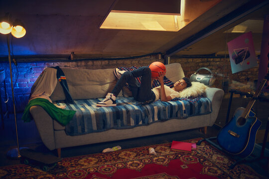 Boy relaxing on sofa with basketball in retro attic room. Concept of leisure, love for sports, and personal space filled with cozy nostalgic atmosphere and youthful energy.