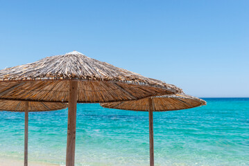 Straw parasols on sandy shore overlooking turquoise sea under bright summer sky, ideal for travel and relaxation themes