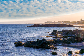 black rock sea beach during sunrise or sunset with golden sand, mild surf, blue calm water and beautiful cloudy sky with coast and mountains on background
