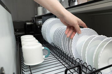 A person is carefully arranging a stack of clean plates with delicate patterns and white mugs in a...